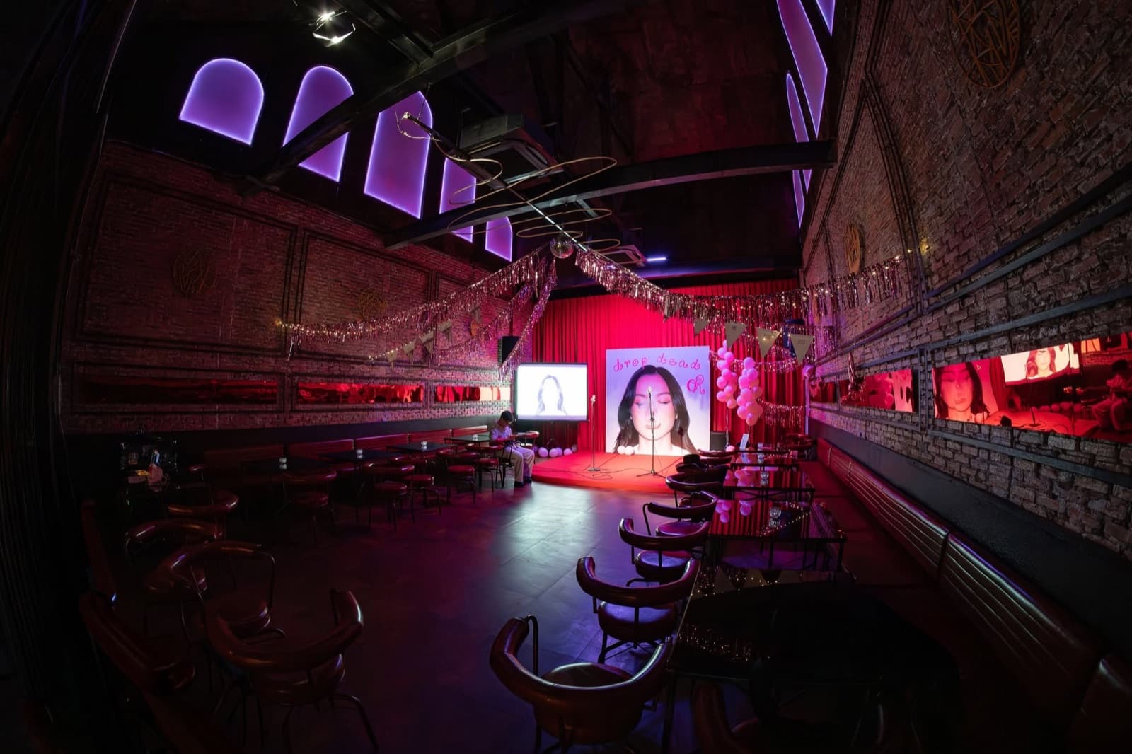 Bebop's main room decorated for an event, red-lit brick walls, a small stage with a lit screen, and rows of leather chairs and tables, unoccupied.