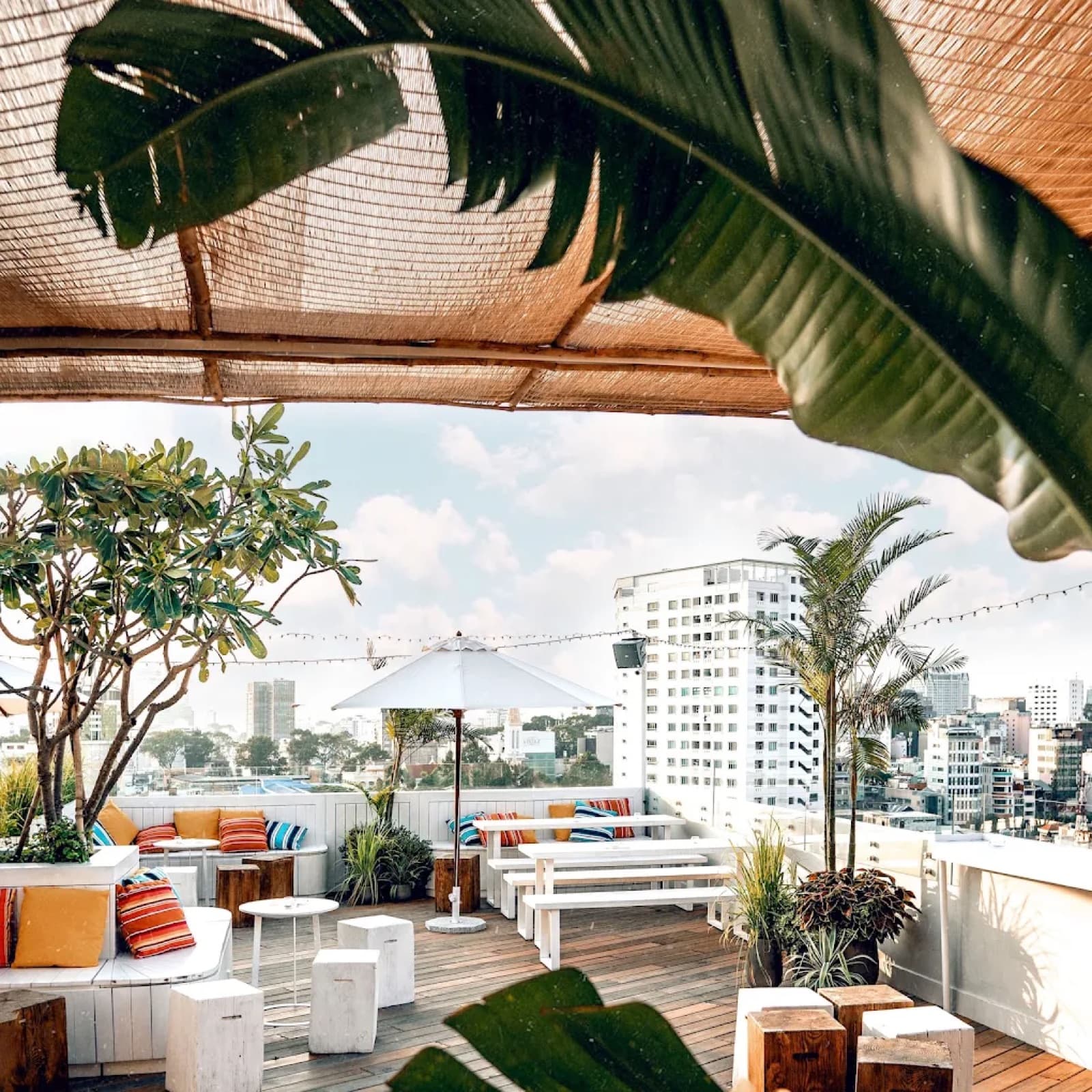 Empty rooftop terrace at Banana Mama with a thatched shade roof, striped lounge cushions and a white umbrella against the Ho Chi Minh City skyline