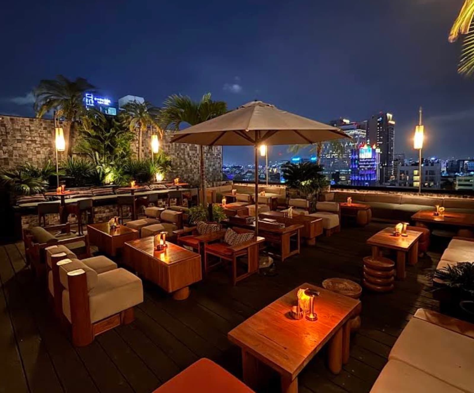 Open-air rooftop terrace at Papaya Papa at night, unoccupied, with low timber tables, banquette seating, parasols and the lit city skyline behind