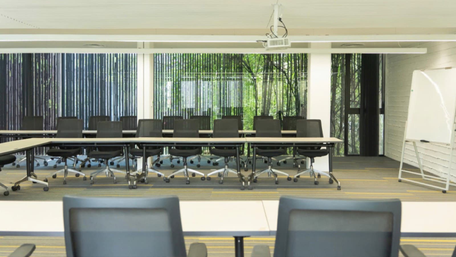 A long boardroom table with black mesh swivel chairs down both sides, a whiteboard at the end and a ceiling projector, with slatted screens and greenery through the windows behind